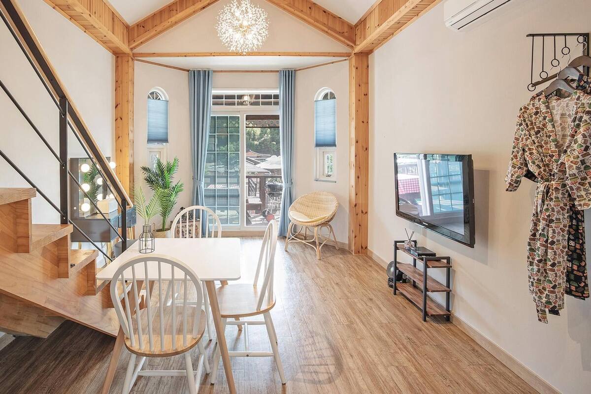 A modern interior with a wooden staircase, a dining area with white chairs, a large window, and a TV mounted on the wall.
