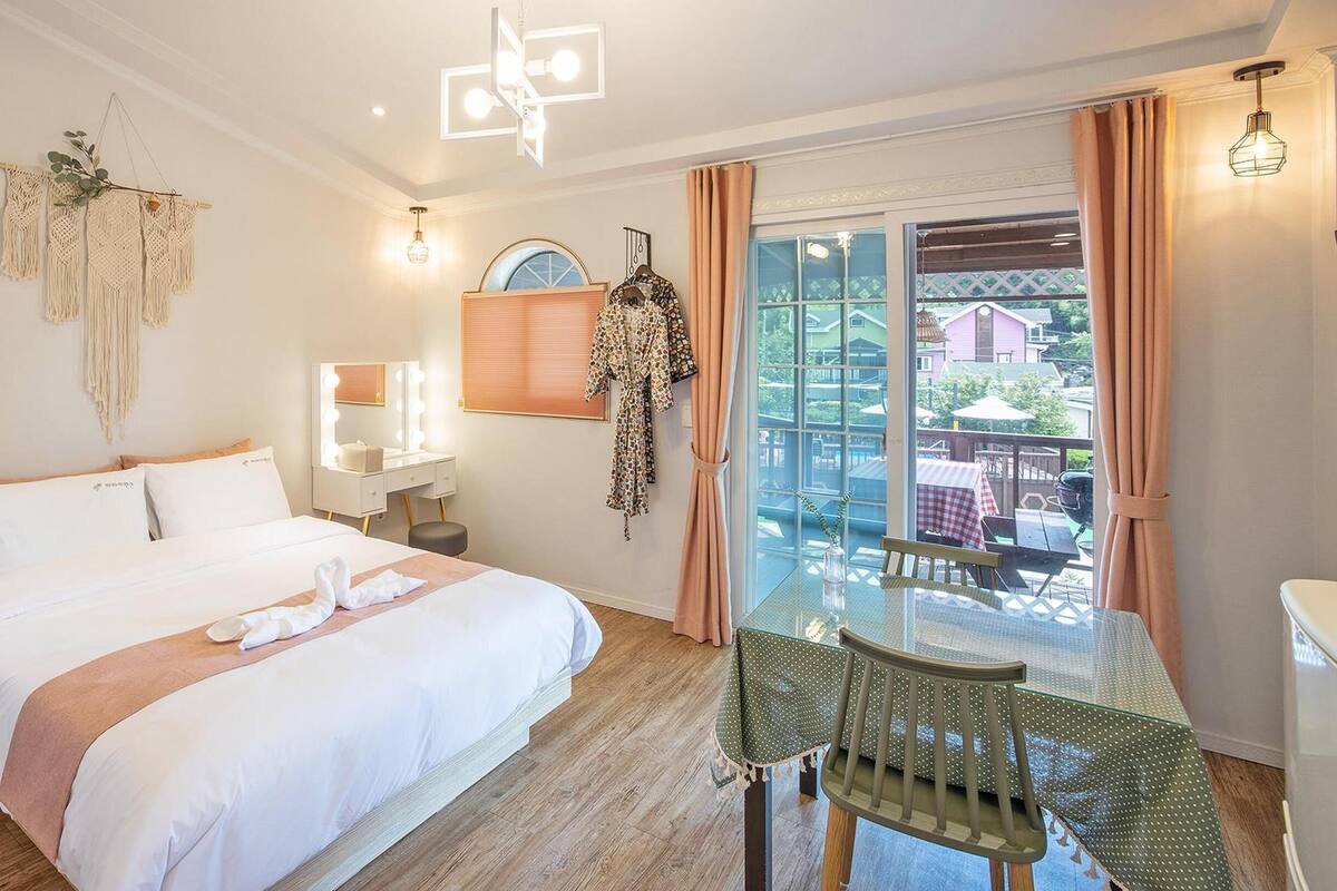 A hotel room with a bed, a vanity with a mirror, a hanging robe, a table with chairs, and a balcony with a view of buildings.