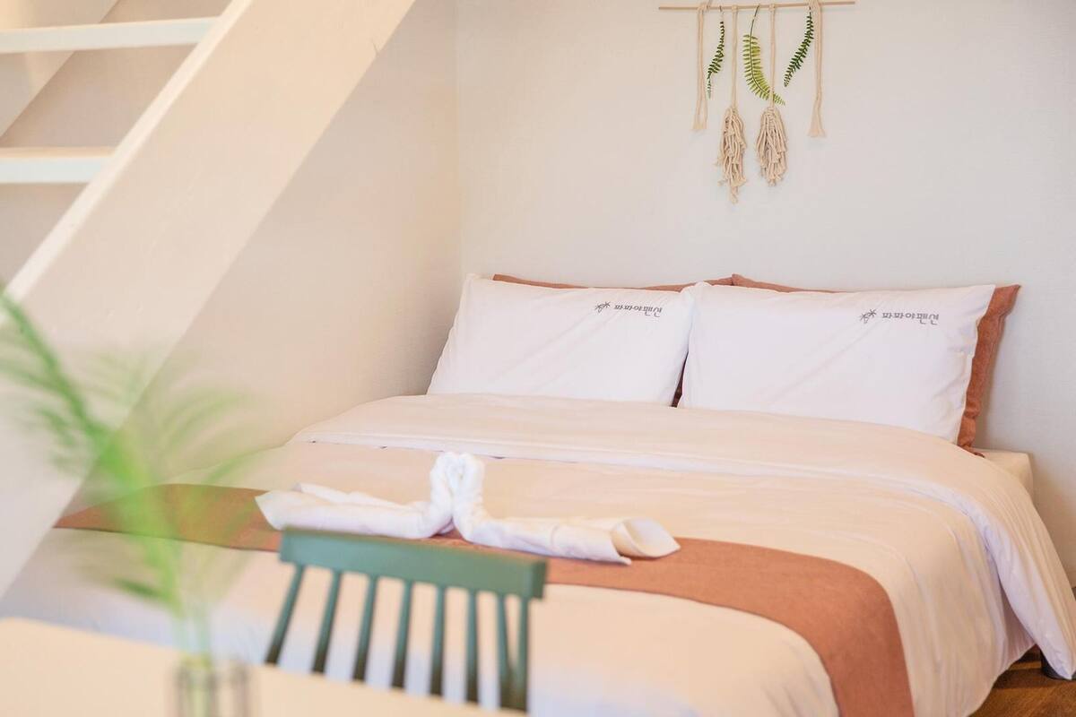 A bedroom with a bed, pillows, a bedside table, a chair, a plant, and a hanging macrame decoration.