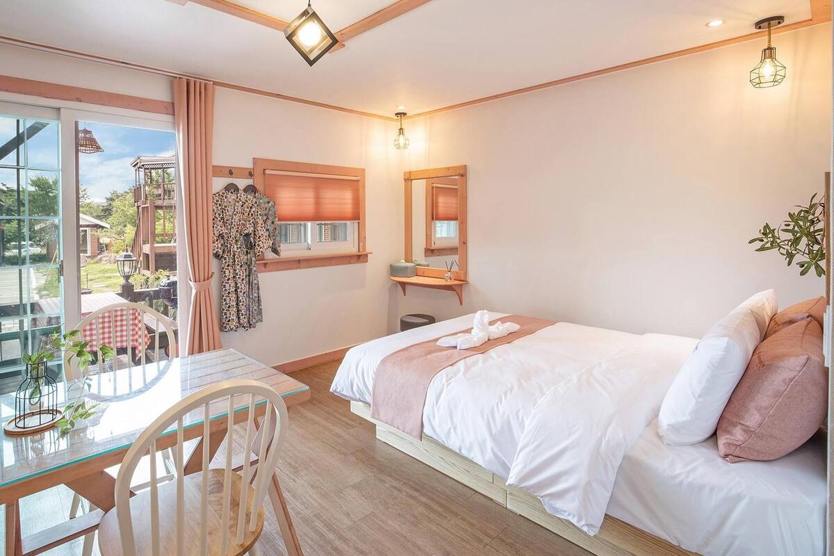 A hotel room with a bed, a table with chairs, a window with curtains, a balcony, and a view of the outside.