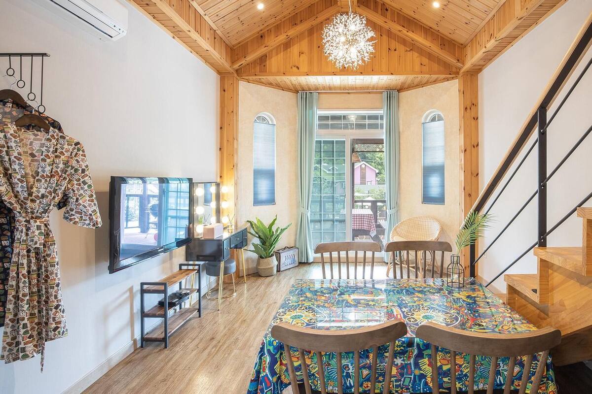 A spacious room with a wooden ceiling, a large mirror with lights, a dining table with colorful tablecloth, and a staircase.