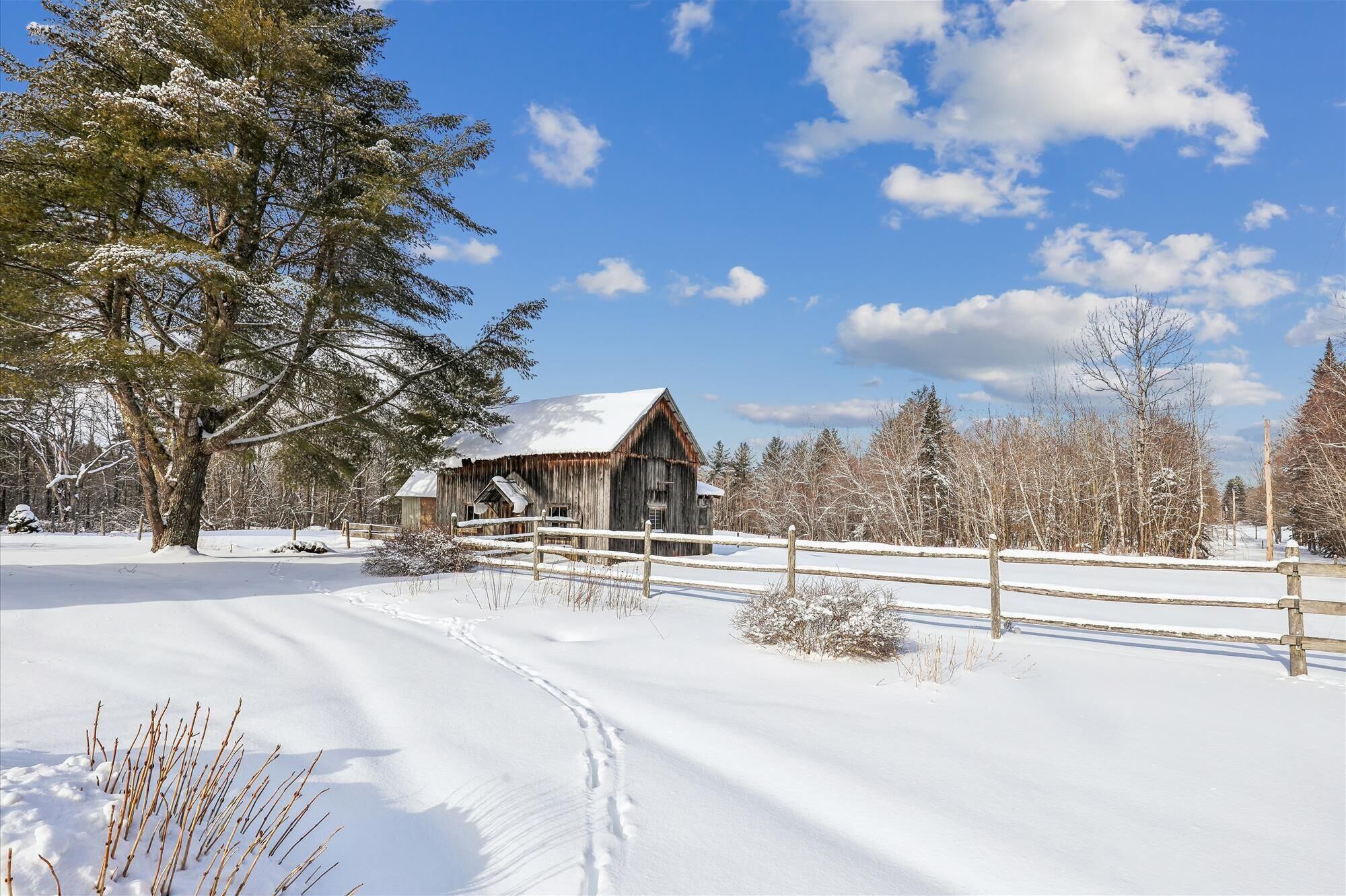 A rustic wooden barn with a snow-covered roof, surrounded by a wooden fence and snow-covered trees.