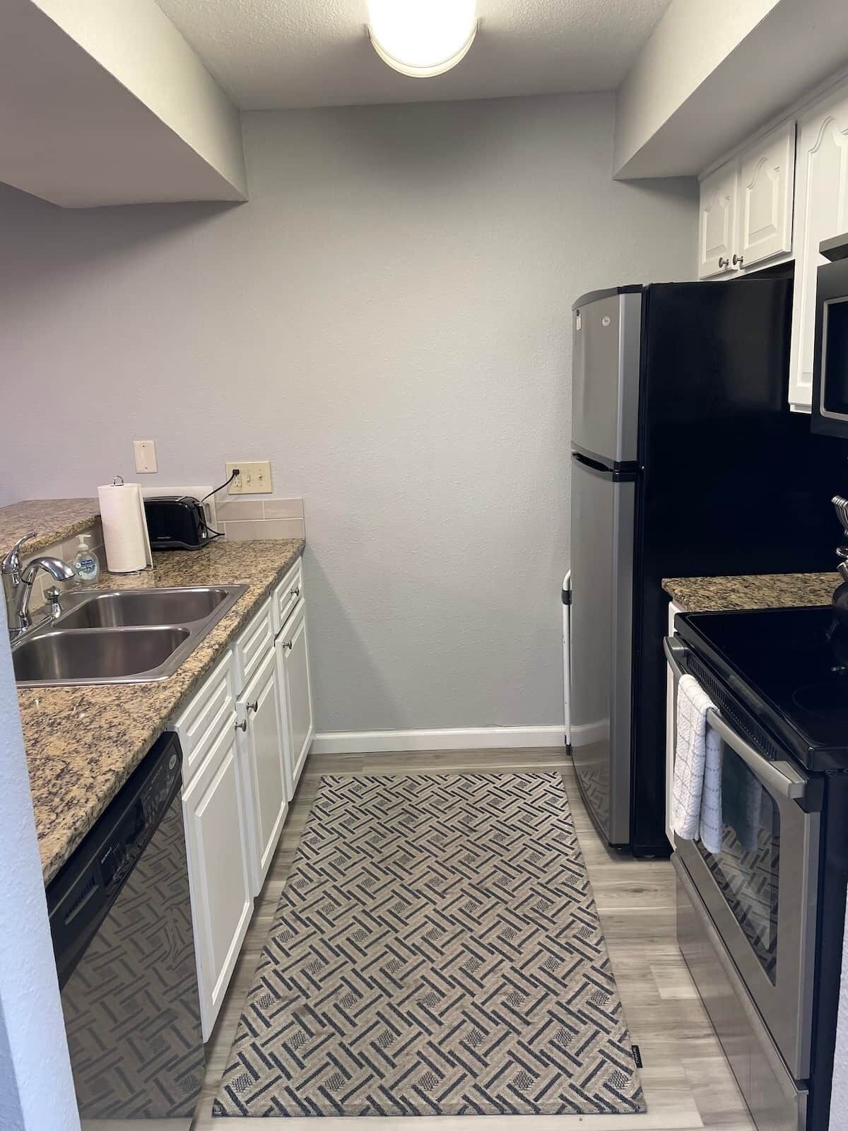 A compact kitchen with a granite countertop, stainless steel sink, white cabinets, a black refrigerator, and a patterned rug.