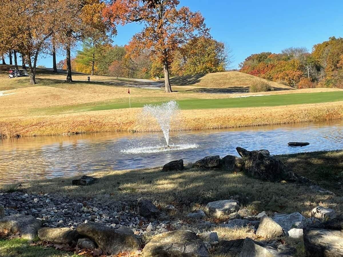 Un étang avec une fontaine, entouré de pelouse et d’arbres aux feuilles d’automne.