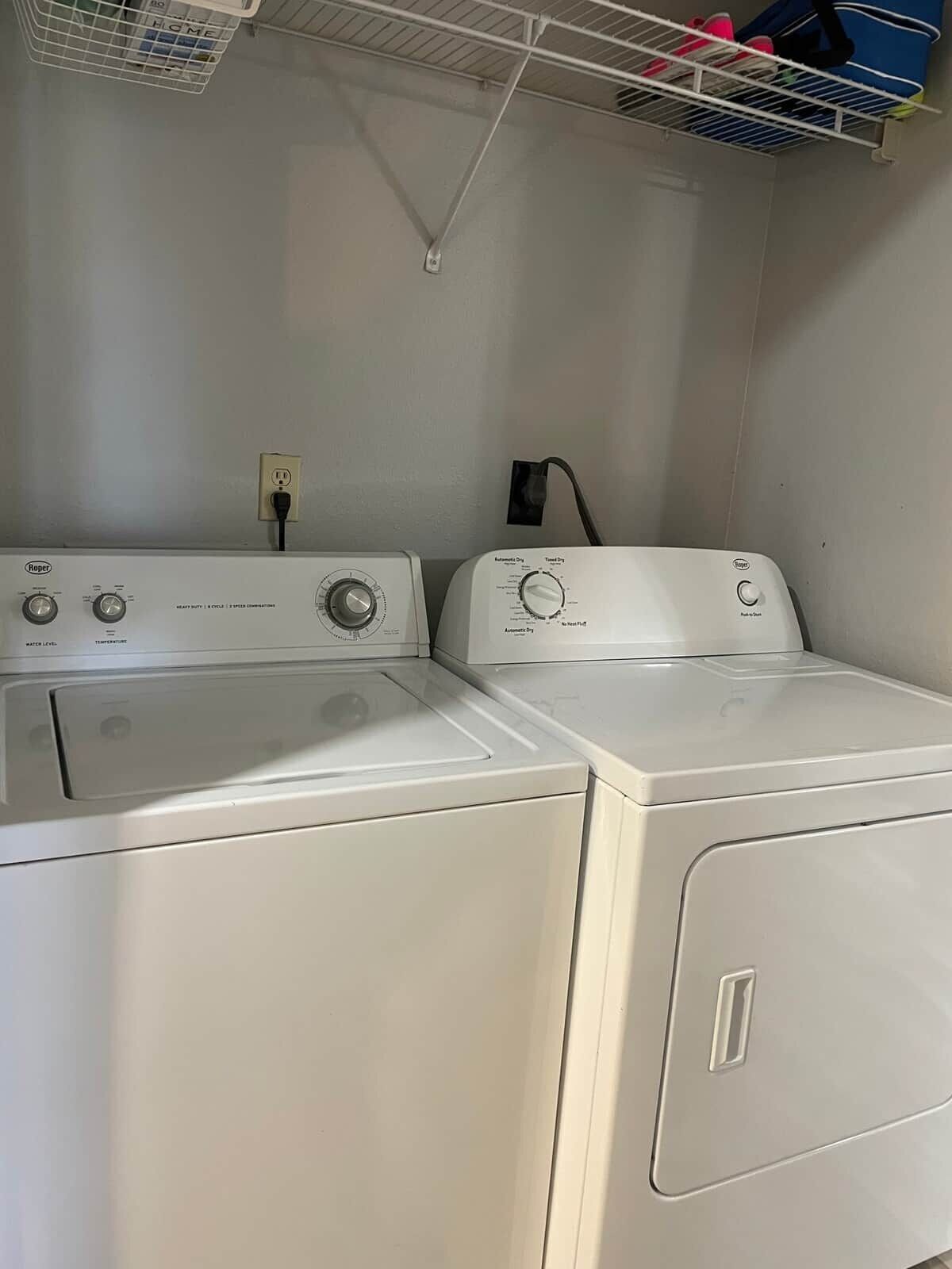 A laundry area with two white front-loading washing machines, a power outlet, and a metal shelf above.