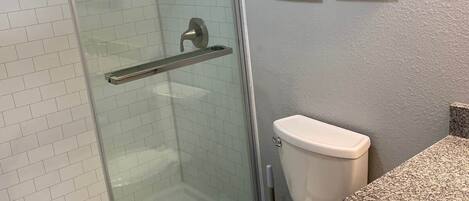 Baño con ducha envuelta en vidrio, inodoro y lavamanos con encimera de granito. Se observan letreros en la pared que dicen 'CEPILLO LAVAR' y 'PASAR HILO DENTAL DESCAGAR'.