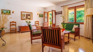 A cozy living room with a wooden sofa, a wooden chair, a small table, and a green cushioned bench. There is a door leading to a garden, and a window with curtains.