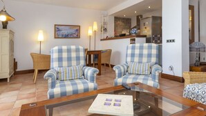 Uma sala de estar com duas poltronas xadrez, uma mesa de centro de vidro, uma cadeira de vime e um quadro na parede.