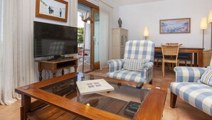 Uma sala de estar com televisão, uma mesa de centro de vidro, uma poltrona xadrez e um espaço de jantar com mesa e cadeiras.