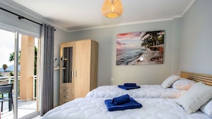 Ein Schlafzimmer mit einem großen Bett, einem Kleiderschrank, einem Bild mit einer Strandansicht und einem Balkon mit Blick auf Palmen.
