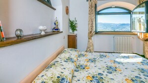 A bedroom with a floral bedspread, a wooden shelf with decorative items, a window with a view of the countryside, and a radiator. - Beautiful apartment for 4 guests with WIFI, pool, A/C, TV, patio and panoramic view (Greve In Chianti)