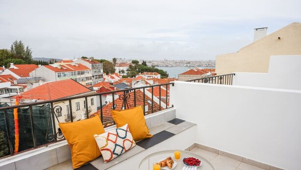 A balcony with a view of a cityscape, a round table with breakfast items, and orange cushions. - One bedroom Apt | Almada | Balcony w/River Views by Lisbeyond (Almada)
