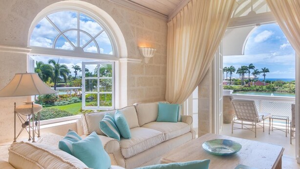 A room with a beige sofa, teal cushions, a glass table, and a view of a landscaped garden and ocean. - Forest Hills 23 | Beach View - Located in  Wonderful Saint James with House Cleaning Included (Saint James)