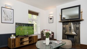 Een woonkamer met een televisie waarop een tenniswedstrijd te zien is, een open haard en een glazen salontafel met een bloemenvaas.