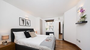 Een moderne slaapkamer met een groot bed, nachtkastje en een bijzettafeltje. Er is een raam met jaloezieën, een deur naar een andere kamer en een plank met een potplant.