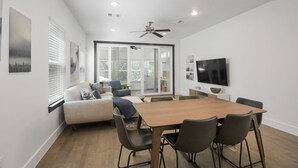 A modern living room with a wooden dining table, comfortable seating, a flat-screen TV, and a ceiling fan.