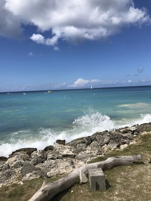 A rocky shoreline with clear turquoise water, a sailboat in the distance, and a clear sky.