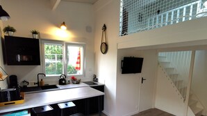 A modern kitchen with a central island, black bar stools, a sink, and a window with a view of greenery. - L'Annexe - Studio 2/4 pers - Private pool (Les Sables-d´Olonne)