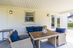 Une terrasse couverte, aménagée avec des meubles en osier et des coussins bleus, une table en bois et deux lanternes fixées au mur.