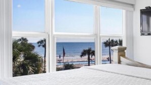 Une chambre avec une grande fenêtre donnant sur une plage, un ventilateur de plafond et un lit recouvert d’une couette blanche.