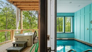 A wooden deck with a hot tub and a small table, a pool with a blue interior, and a view of trees through a window.