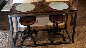 Una mesa de madera con dos taburetes, un jarrón con plumas de plumas de pampa secas y un adorno metálico en la pared.