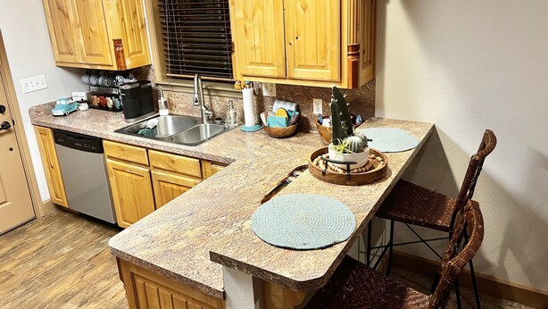 Una cocina con armarios de madera, encimera de granito, lavavajillas, pileta y comedor con sillas de mimbre.