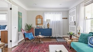 Una sala de estar con un sofá azul, una mesa de centro, una silla, un armario azul, un espejo y una ventana con cortinas.