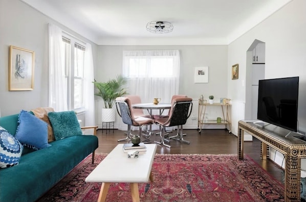 A living room with a green sofa, a white coffee table, a dining table with chairs, a TV, and a large window with curtains.