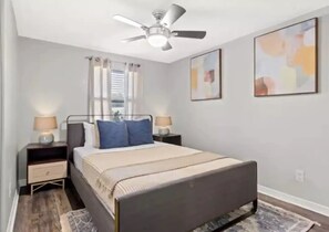 A bedroom with a bed, two bedside tables, a ceiling fan, a window with curtains, and two abstract paintings on the wall. - Newly renovated outdoor space, hot tub, fire pit,minutes from the beach. (Largo)