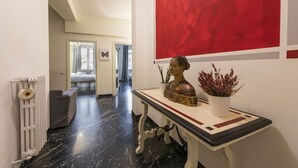 A hallway with a radiator, a grey sofa, a table with a bust sculpture and a potted plant, and a red abstract painting on the wall.
