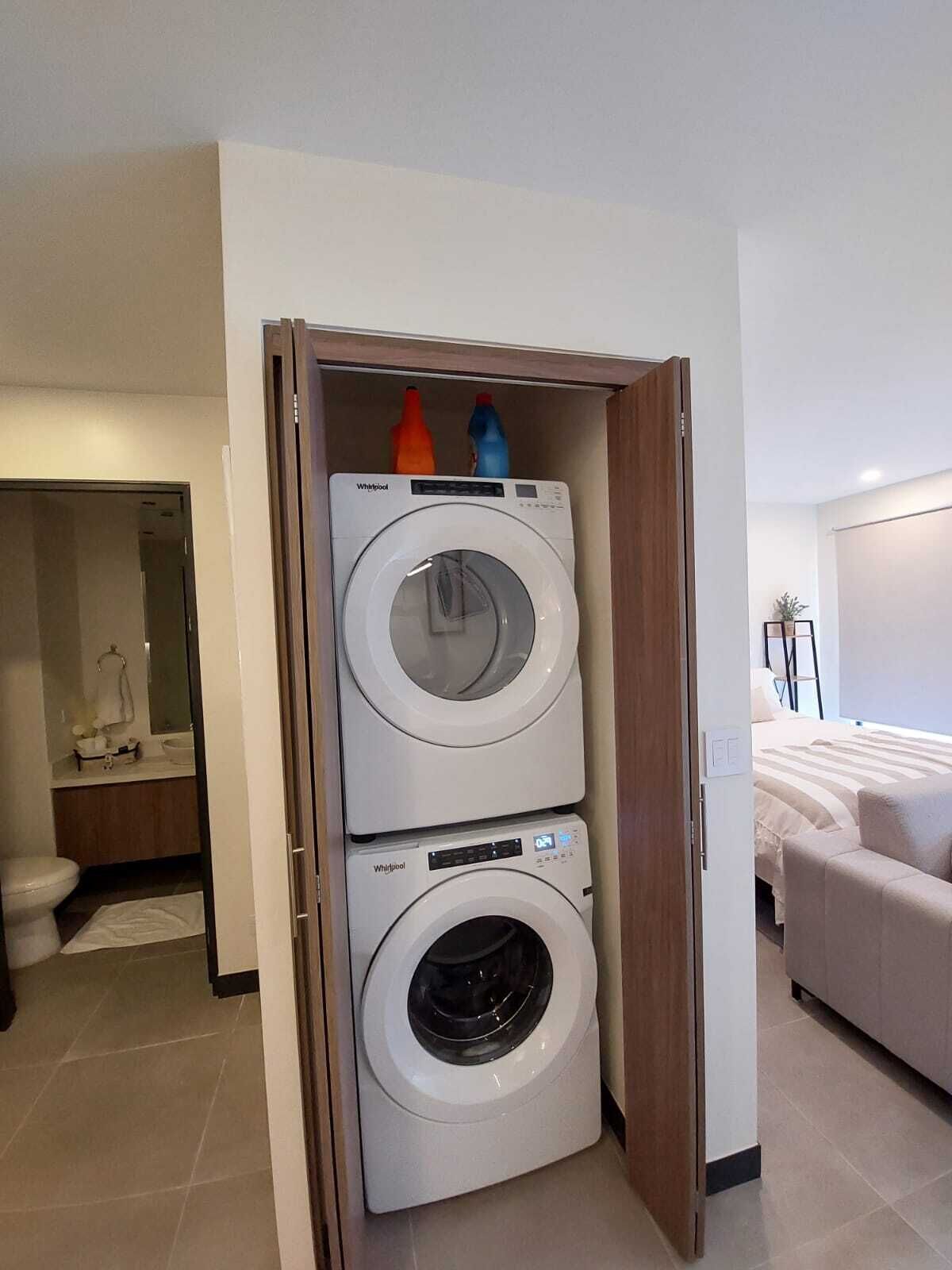 Een praktische wasruimte met twee gestapelde Whirlpool wasmachines en drogers, een zichtbare badkamer met toilet en wastafel, en een bed in de aangrenzende kamer.