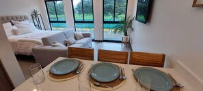 Une chambre d’hôtel moderne, équipée d’un lit, d’un canapé, d’une table à manger pour quatre personnes et de grandes fenêtres donnant sur l’extérieur.