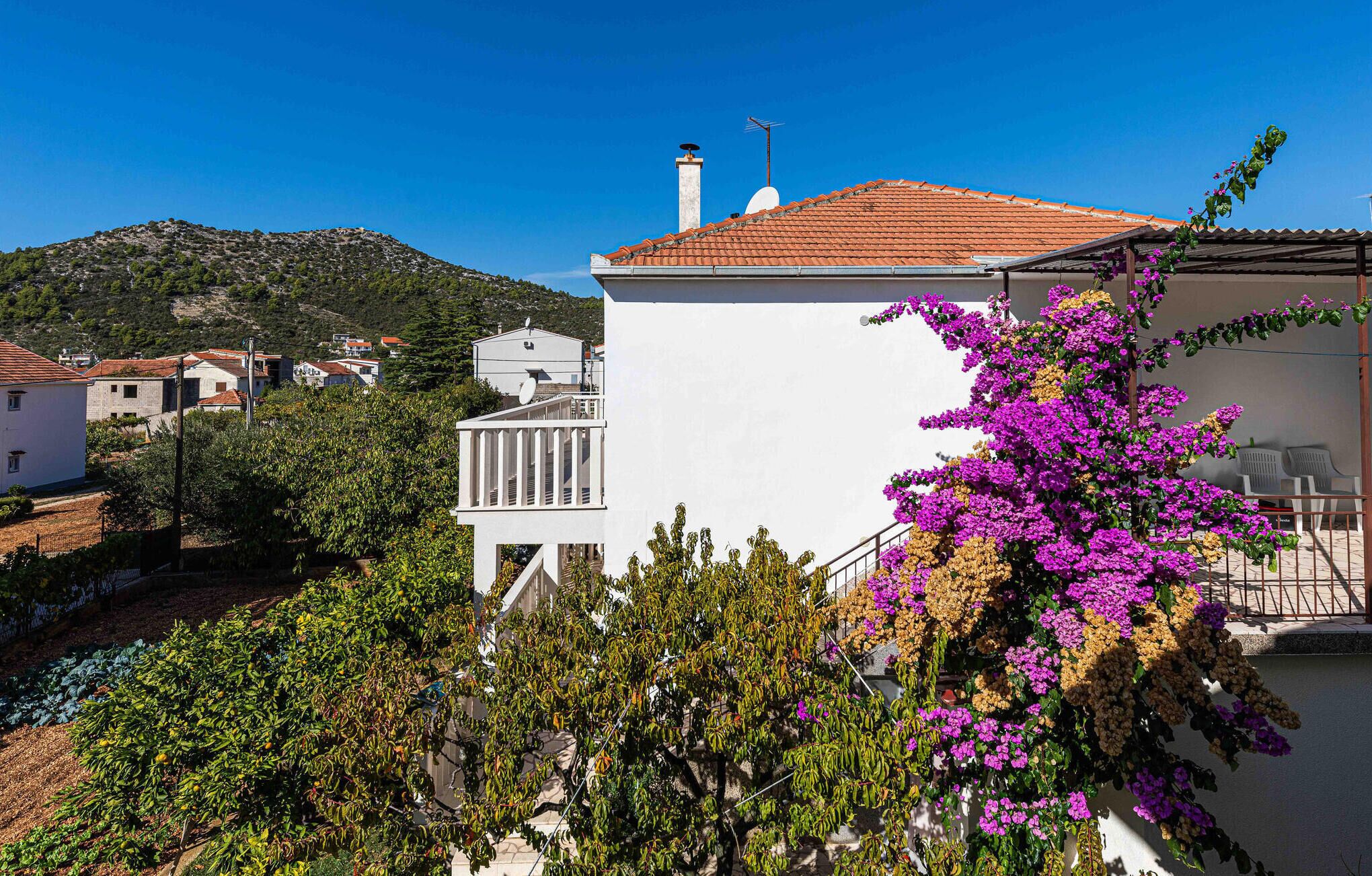 A white building with a red-tiled roof, surrounded by greenery and a mountainous backdrop.
