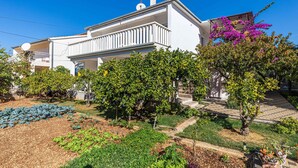 Ein weißes, zweistöckiges Haus mit Balkon, umgeben von einem Garten mit verschiedenen Pflanzen und einer Pergola mit violetten Blüten.
