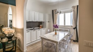 A modern kitchen with white cabinets, a white dining table with clear chairs, and a window with curtains. - Apartments Florence - Borgo dei Greci (Firenze)