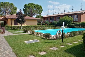 Un área de piscina con una piscina azul cristalina, rodeada de césped y setos bien cuidados. Se observan dos edificios con balcones y antenas parabólicas.