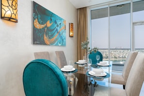 A dining area with a glass table, teal chairs, and a city view through large windows.