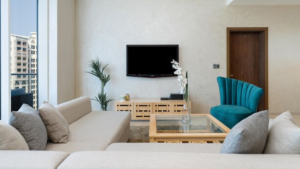 A modern living room with a beige sofa, a teal armchair, a glass coffee table, and a flat-screen TV mounted on the wall. - Modern, Charming, Sea View Apt. In Palm Jumeirah (Dubai)
