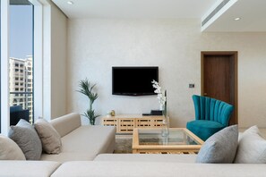 A modern living room with a beige sofa, a teal armchair, a glass coffee table, and a flat-screen TV mounted on the wall.