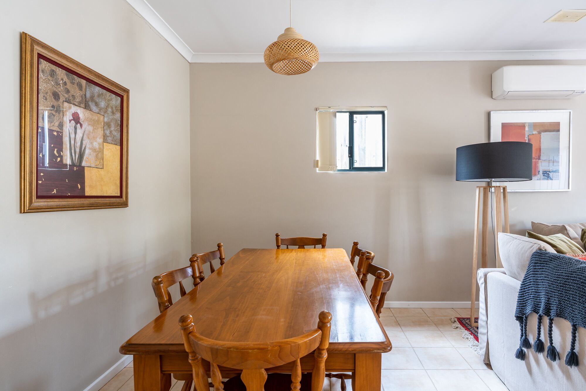 A dining area with a wooden table and chairs, a hanging light fixture, a framed artwork, and a window with blinds.