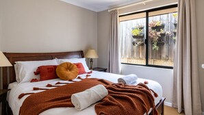 A neatly made bed with a brown knitted blanket and pillows, a bedside lamp, and a window with curtains.
