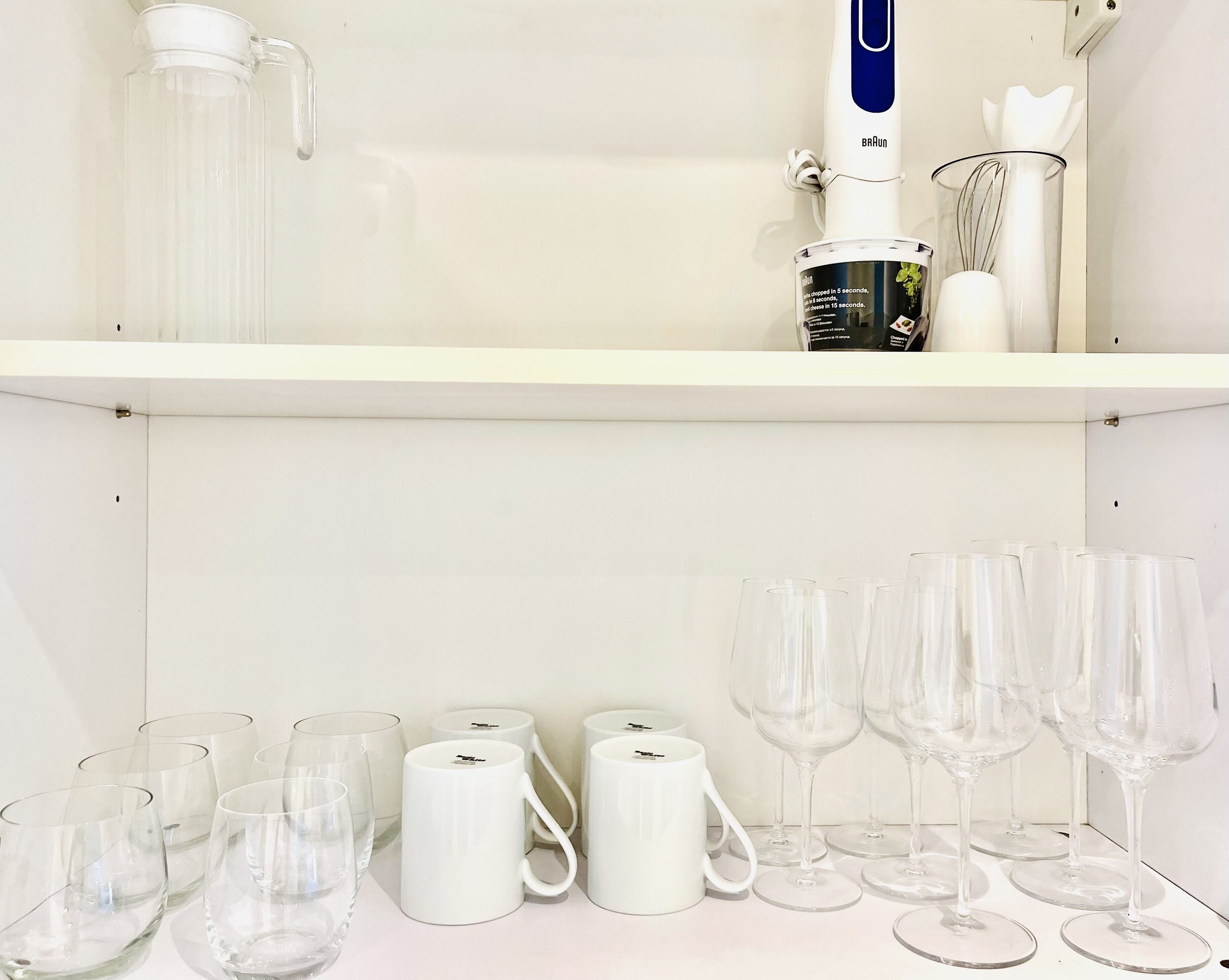 A kitchen area with a Braun coffee maker, a glass pitcher, and a coffee machine on the shelf. Below, there are several wine glasses and a white mug.