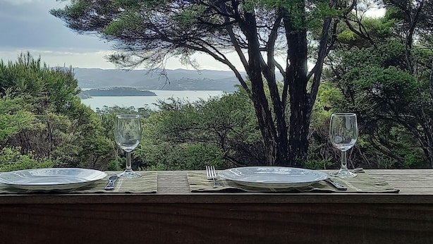 A table set with plates, cutlery, and wine glasses, overlooking a scenic view of a lake and mountains. - Te Oneroa Hill, 5 acres of native park with wonderful sea view. (Russell)