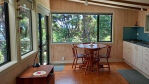 A kitchen with wooden cabinets, a dining table with chairs, and large windows offering a view of trees. - Te Oneroa Hill, 5 acres of native park with wonderful sea view. (Russell)
