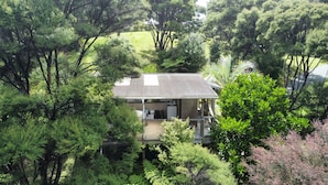 A house surrounded by dense trees, with a visible garage and a covered porch. - Te Oneroa Hill, 5 acres of native park with wonderful sea view. (Russell)