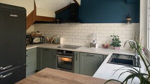 Een moderne keuken met een zwarte koelkast, een magnetron, een broodrooster en een blender op het aanrecht. Er staat een houten tafel en een gootsteen met een potplant ernaast.