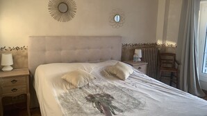 Slaapkamer met een knopen hoofdbord, twee nachtkastjes met lampen, een radiator, een houten stoel en een raam met gordijnen.