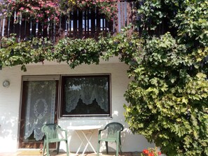 Ein Balkon mit Holzgeländer, einem Tisch mit Stühlen und einem Fenster mit Spitzenvorhängen.