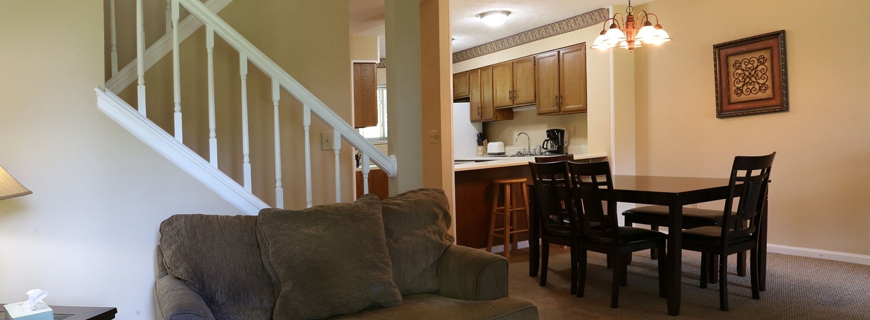 A living room with a sofa, a dining table with chairs, a kitchen area with wooden cabinets, and a staircase.